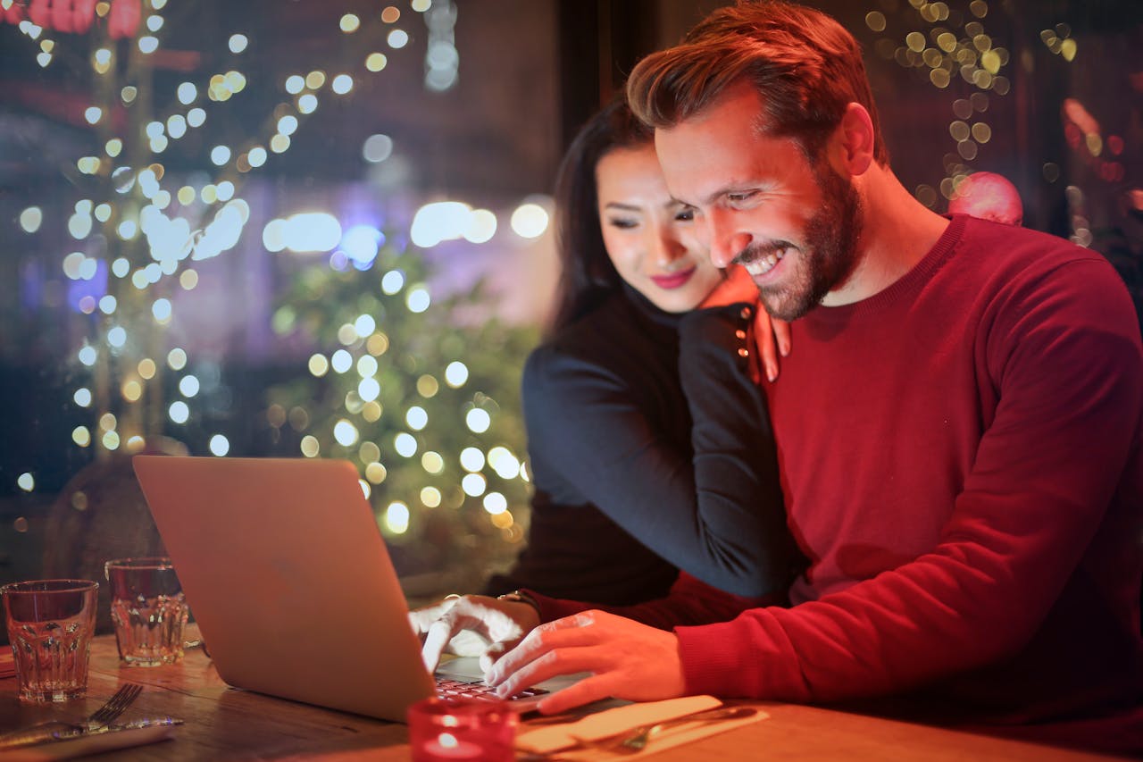 home-img A couple using a laptop at a cozy restaurant with festive bokeh lights, enjoying online shopping together.